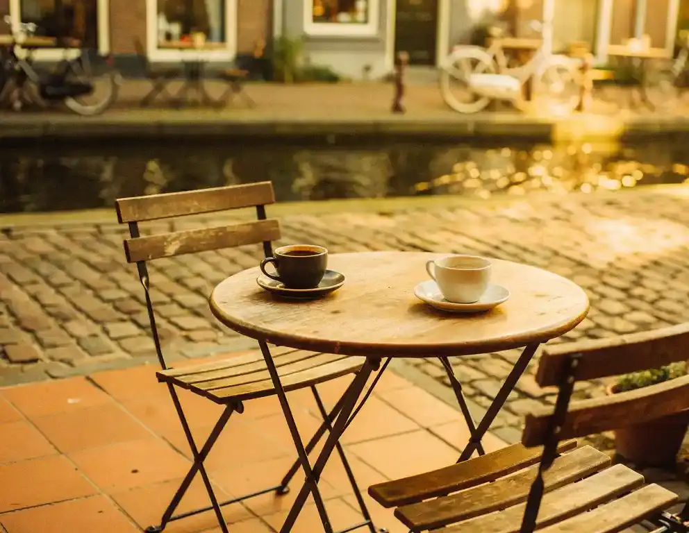 Lege stoel aan terrastafeltje in Amsterdam met halfvol koffiekopje, zacht kanaallicht op kasseien op de achtergrond.