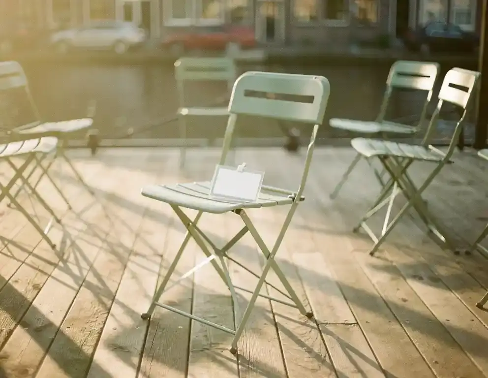 Lege klapstoel met vergeten naambordje, iets apart van een kring stoelen op een Amsterdams kanaalterras in middagzon.