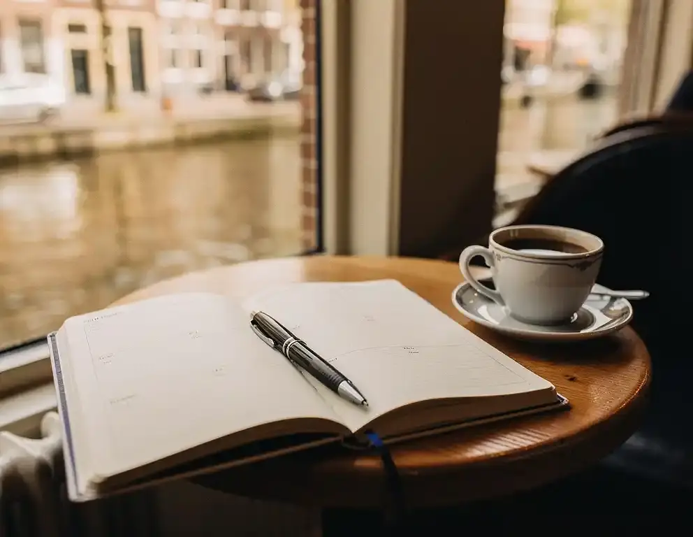 Open notitieboek met handgeschreven planning op houten cafétafel, pen ernaast, koffie en kanaalgracht op de achtergrond.